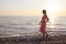 Young woman in pink dress slowly walks along the coastline of surf barefoot and looks at horizon