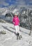 Young woman in pink jacket, ski, boots, poles, gloves and hat, posing on the piste, snow covered mountain behind her during sunny