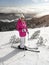 Young woman in pink jacket, skis, ski poles, goggles and hat, posing on the snow covered piste, sun shining to forest behind her
