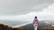 Young woman in pink jacket thoughtful walk towards edge cliff viewpoint watch atlantic ocean waves in Iceland.Black sand beach