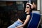 Young woman with pink maroon burgundy short hair, wearing black top and light blue jeans, sitting on coach in subway train. Three-