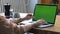 Young woman in pink shirt sitting at the large wooden table and typing on her laptop with chroma key green screen. Stock