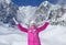 Young woman in pink ski jacket, gloves and winter hat, smiling, throwing snow in the air, sun shining on mountain behind her.