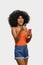 Young woman with positively surprised afro hair points to a white space, she holds a smart phone in one hand,