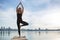 Young woman practicing yoga exercise at quiet wooden pier with city background. Sport and recreation in city rush