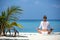 Young woman practicing yoga meditation on the beach facing the ocean near a palm tree on Maldives