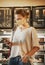 A young woman in a protective mask stands in a cafe at the counter with desserts and a cash register with a smartphone in her