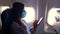 Young woman in a protective mask uses a mobile, smartphone inside airplane, sitting near an illuminator. Resumption of