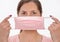 Young woman putting pink cotton home made virus mouth face mask on, holding it in front of her, portrait on white background, can