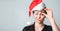Young woman raised glasses, squint and looks at the camera in santa claus hat. Doubtful and perplexed look. Bright shocked