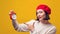 Young woman in red beret with alarm clock. Woman pointing on clock over yellow background. Girl shows the time on the clock.