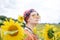 Young woman with red burgundy hair, wearing boho hippie clothes and yellow sunglasses, standing in middle of sunflowers field.