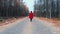 Young woman in red coat walking alone along empty road in autumn forest. Back view. Travel, freedom, nature concept