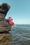 A young woman in a red dress and white hat sits on a rock by the sea and holds a hat with her hands. Clear sky