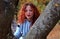 A young woman with red hair attentively looks into the camera and clings to a tree in the park. on the background you can see the