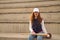 Young woman with red hair, freckles, wearing white cap, jacket, baseball bat and glove, sitting on concrete bleachers. Concept