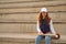 Young woman with red hair, freckles, wearing white cap, jacket, baseball bat and glove, sitting on concrete bleachers. Concept