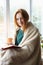 young woman with red hair reading book and drinking coffee wrapped in soft blanket