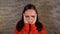 A young woman in a red hoodie grabs her head with hands in pain on a brick background. A woman has a headache.