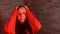A young woman in a red hoodie grabs her head with hands in pain on a brick background. A woman has a headache.