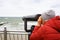 A young woman in a red jacket and a knitted cap looks with interest through a telescope at the sea with waves, standing on the