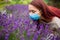 Young woman with red long hair wearing surgical mask trying to smell flowers of purple lavender. Allergy, covid, flu.
