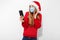 Young woman in a red Santa Claus hat and a medical mask on her face, waving, making a video call on a smartphone on Christmas eve