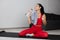 Young woman in red tracksuit doing exercise or yoga at home. Sitting alone on mat and posing with water bottle. Drinking