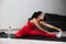 Young woman in red tracksuit doing exercise or yoga at home. Well-built strong sporty girl stretching her left leg and