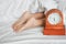 Young woman`s feet under white blanket on bed and blurred clock