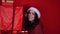 Young woman in Santa Claus hat holds big gifts, standing near red wall. Happy female in Christmas hat with boxes of gift