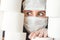 Young woman with scared eyes in two medical virus protection face masks looks through stacks of toilet paper. Covid-19 quarantine