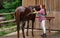 Young woman in shirt washing brown horse after ride, smiling, water flowing from hose near stables