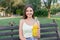 Young woman is showing a bottle of cool juice on the bench in the park