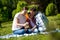 Young Woman Is Showing Photos On Phone To Her Husband And Daughter While Sitting On The Blanket In The Park.