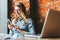 Young woman sits in cafe on windowsill and uses smartphone. Hipster girl checking e-mail, chatting, blogging, working.