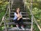 Young woman sits on old wooden staircase in woodland. Adult brunette poses, looking at camera and resting on stairs in