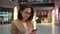 A young woman sits with a phone in her hands in a shopping center with a large paln. A girl in a pink coat is texting on
