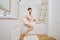 Young woman sitting on bathtub applying body lotion, touching soft smooth silky legs in bathroom