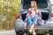 Young woman sitting on car trunk with coffee cups and thermos on the side of the road in the forest in warm fall or