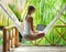 Young woman sitting in a hammock with laptop in a tropical resort. back view.