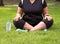 young woman sitting have plastic bottle beside her on grass field in public garden for meditation