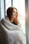 Young woman sitting near the window wrapped in blanket, relaxing, thinking, warming up, cold autumn morning, lazy day off