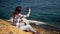 A young woman is sitting with a phone on the stones in front of the sea. Girl in sunglasses herself. Splashes of sea