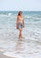 Young woman in skirt, t shirt and sunglasses standing in shallow water on the beach during overcast day, sea behind her