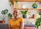 A young woman smiles while sitting on the couch- bookshelf filled with plants