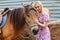 Young woman smiling and stroking a brown horse before a walk that eats hay near the haystack on a summer clear day