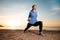 A young woman in sportswear trains by performing a leg stretch. In the background, sunset, sandy beach and ocean. Recreation and