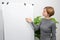 A young woman is standing in a bright room pointing at a blank flip chart. Online teacher, tutor