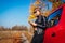 Young woman standing by car using smartphone on autumn road. Driver stopped auto to catch better signal